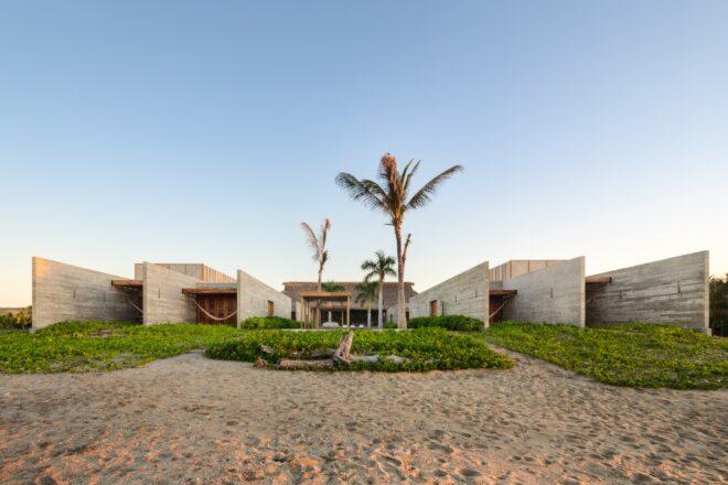 The exterior of a beach house, with sand, greenery and palm trees in the front yard. The home is comprised of a U-shaped, off-white wood volume.