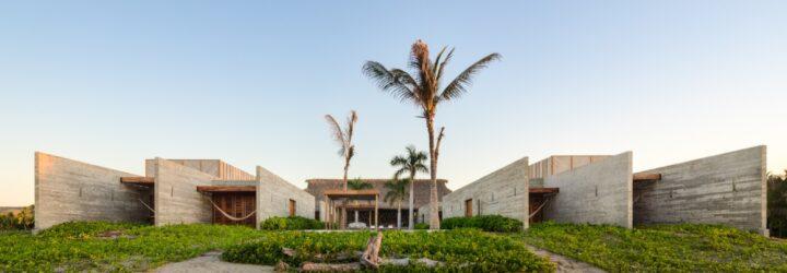 The exterior of a beach house, with sand, greenery and palm trees in the front yard. The home is comprised of a U-shaped, off-white wood volume.