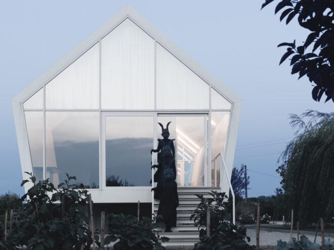 a pentagon-shaped house with white frames and a glass facade. three people in all black with black rabbit heads on stand on the stands.