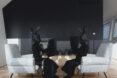 two people in all black with black rabbit heads sit in a living room with white armchairs and a dark accent wall.
