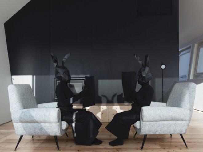 two people in all black with black rabbit heads sit in a living room with white armchairs and a dark accent wall.