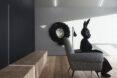 a person in all black with a black rabbit head sits in a living room with a white armchair. a wreath hangs on the wall, with a human face in the middle.
