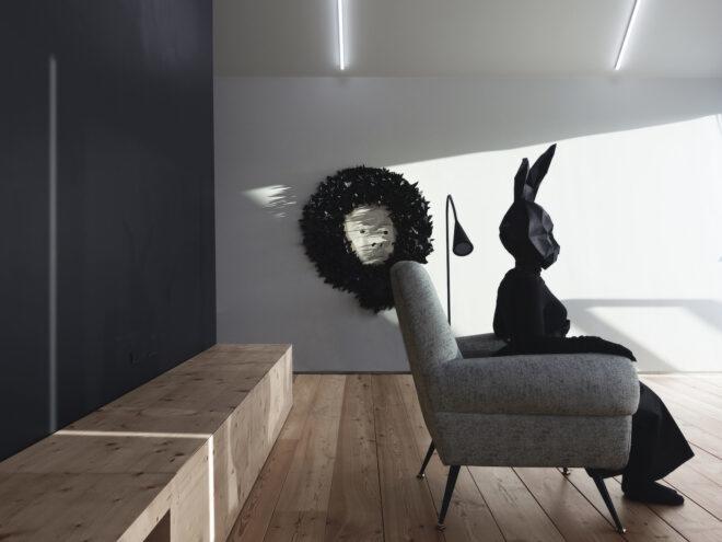a person in all black with a black rabbit head sits in a living room with a white armchair. a wreath hangs on the wall, with a human face in the middle.