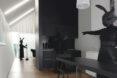 a person in all black with a black rabbit head stands behind a black table, pointing to another person in all black holding a broom next to a set of black cabinets. in the background, another person in all black sweeps the hallway.