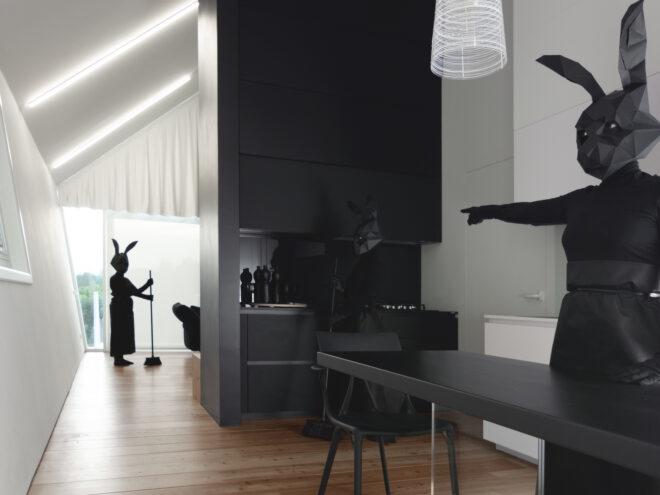a person in all black with a black rabbit head stands behind a black table, pointing to another person in all black holding a broom next to a set of black cabinets. in the background, another person in all black sweeps the hallway.