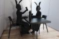 two people in all black with black rabbit heads sit at a black table with black chairs, cups and pitcher.