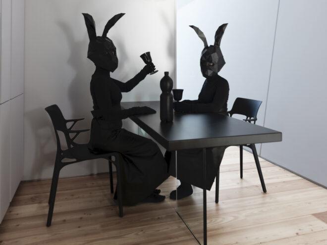 two people in all black with black rabbit heads sit at a black table with black chairs, cups and pitcher.