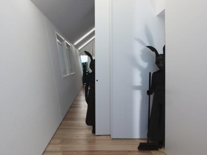 a person in all black with a black rabbit head stands next to a white wall, holding a broom. to the left, another figure in all black hides halfway behind a white door. in the background, a third figure in all black stands in the hallway.