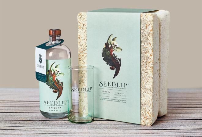 An off-white mycelium box with a blue label reading "Seedlip." Next to the box is a bottle of Seedlip Spice 94 aromatic and a highball glass.