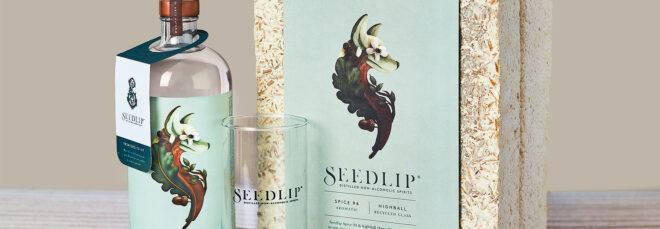 An off-white mycelium box with a blue label reading "Seedlip." Next to the box is a bottle of Seedlip Spice 94 aromatic and a highball glass.