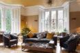 A sitting room with yellow walls, large bright windows, a Christmas tree in the corner, a black sofa and two chairs in front of a rectangular coffee table.