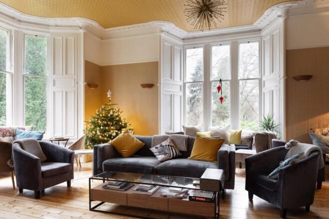 A sitting room with yellow walls, large bright windows, a Christmas tree in the corner, a black sofa and two chairs in front of a rectangular coffee table.