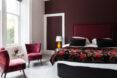 A bedroom with dark mauve walls and a large window letting in bright natural light. Two crimson patterned armchairs sit next to a black and crimson patterned bedspread.