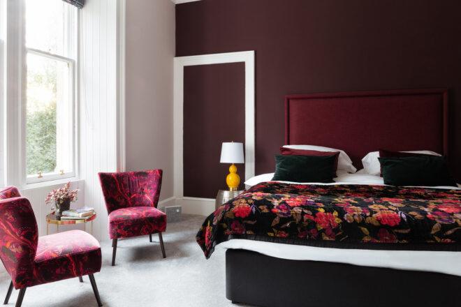 A bedroom with dark mauve walls and a large window letting in bright natural light. Two crimson patterned armchairs sit next to a black and crimson patterned bedspread.