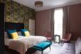 A bedroom with light-colored mauve walls and one accent wall with green patterned wall paper. A blue bench and pillows accent the room.
