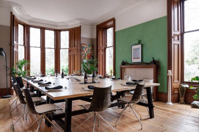 A dining room with a large rectangular wood table surrounded by chairs. The walls are painted green, with large windows toward one end of the room.