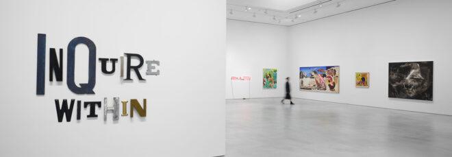 A white gallery space. To the left, a sign that says "Inquire Within." To the right, a blurry figure walks past several hanging art pieces.
