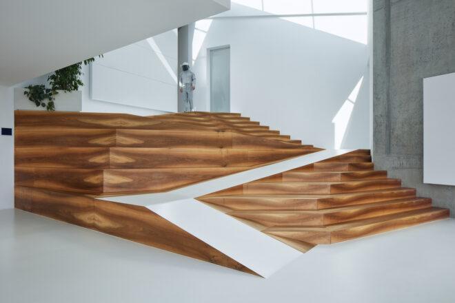 The interior of a home with white walls and floors and a wood staircase with a person in an astronaut outfit standing at the top.