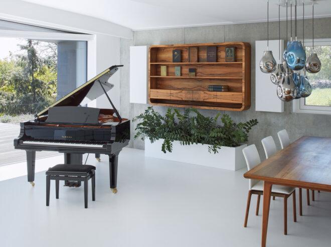 A living area with a bookshelf on the wall, a dark colored piano to the left and a dining table with chairs to the right.