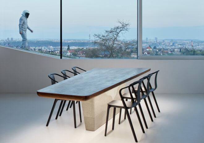 A dining table and chairs sit in front of a long window, where a person in an astronaut outfit walks by outside.