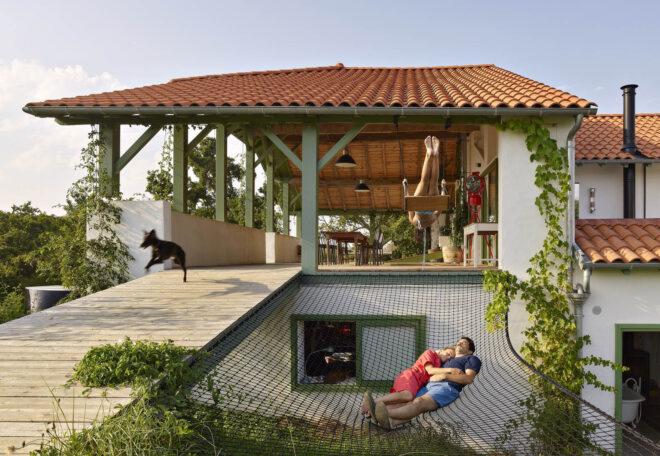 The exterior of a renovated farmhouse with a couple laying on a hammock outside.