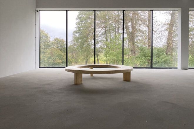 A wood Circula bench in an empty room in front of windows.