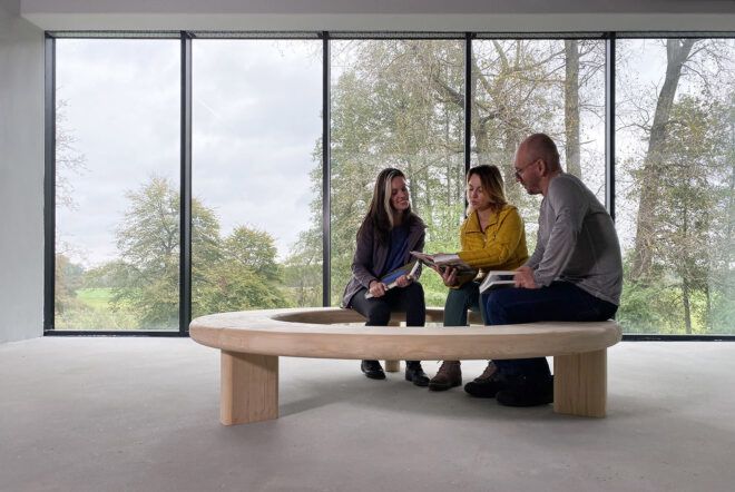 People sitting on a wood Circula bench in an empty room in front of windows.