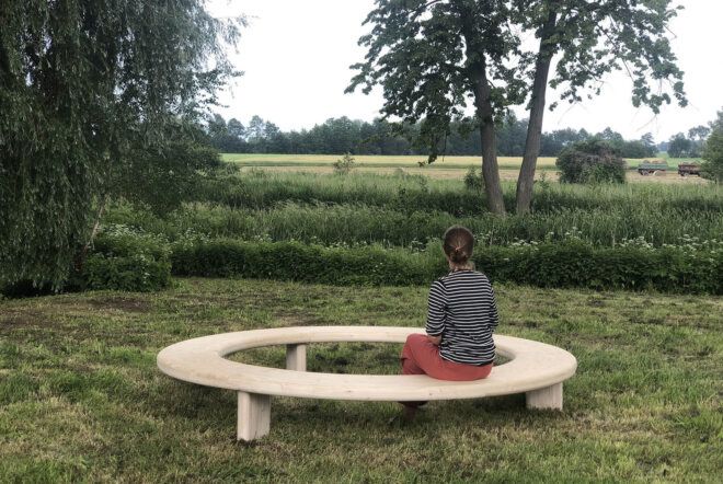 A person sitting on the wood Circula bench out in a grassy area.