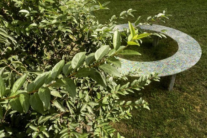 A plastic Circula bench outside in the grass, partially obscured by leaves.