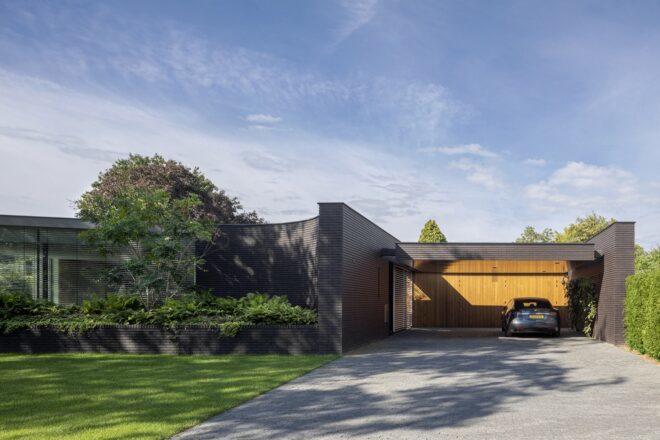 A sleek one-story home with glazing and black bricks covering the facade. To the right is a garage with wood paneling and a car parked inside. In front of the home is a lush green lawn and garden.