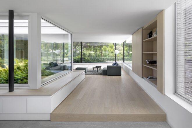 A living room area with white walls and light-colored wood accents. Large windows all around let natural light into an area with a gray sofa and ottoman.