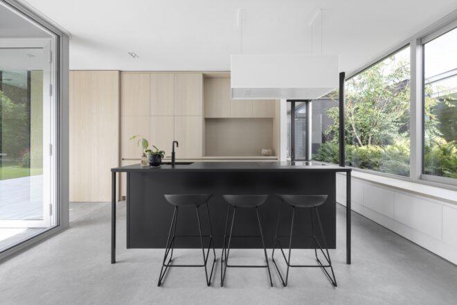 A kitchen flanked on either side by large windows looking out onto greenery. The kitchen itself has a black island countertop with three black stools and a built-in sink. In the background are light-colored wood cabinets.