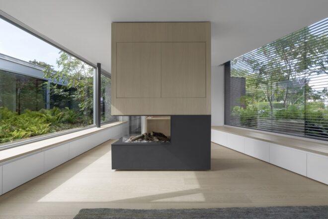 A standalone fireplace with a gray block volume on the bottom and light-colored wood cabinets on top. The fireplace is flanked on either side by large windows that look out onto greenery.