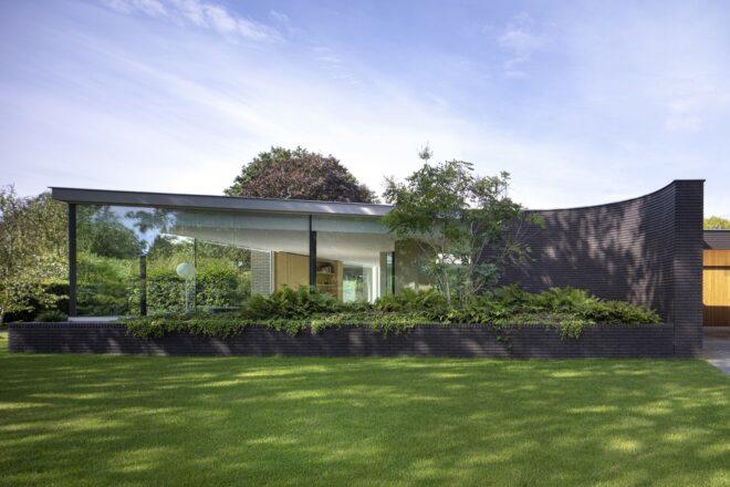 A sleek one-story home with glazing and black bricks covering the facade. In front of the home is a lush green lawn and garden.