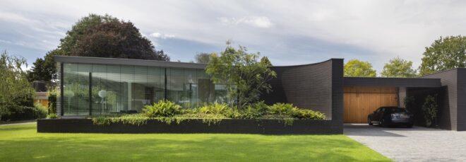 A sleek one-story home with glazing and black bricks covering the facade. To the right is a garage with wood paneling and a car parked inside. In front of the home is a lush green lawn and garden.