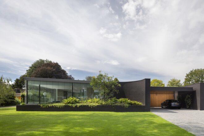 A sleek one-story home with glazing and black bricks covering the facade. To the right is a garage with wood paneling and a car parked inside. In front of the home is a lush green lawn and garden.