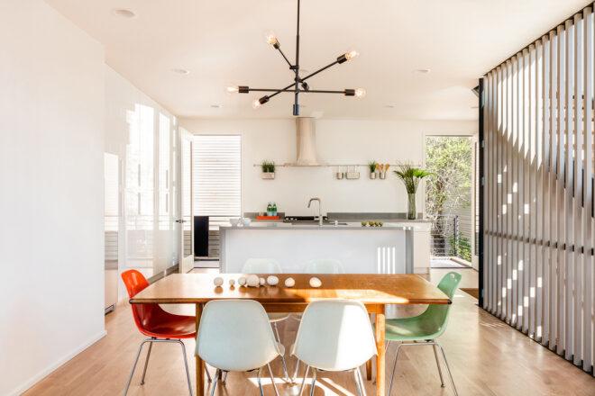 A kitchen area with a rectangular wood dining table and six chairs in the colors white, orange and green. In the background, there is a kitchen with white countertops.