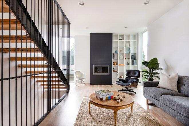 A living room with a stairwell to the left and a gray sofa on the right. In the middle of the room are two chairs, one white and one black, a round wood coffee table and a fireplace.
