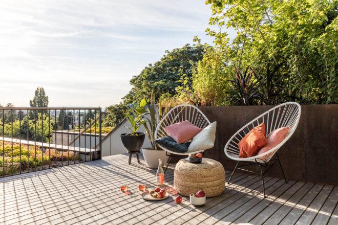 A patio area with two white chairs and a small ottoman surrounded with apples and wine. Greenery surrounds the patio area.