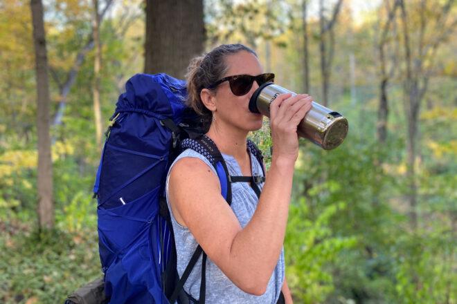 person drinking from metal travel mug while walking in the woods