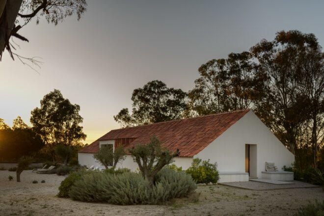 A dusk-time view of a simple A-line house with a red roof, surrounded by trees and green bushes.
