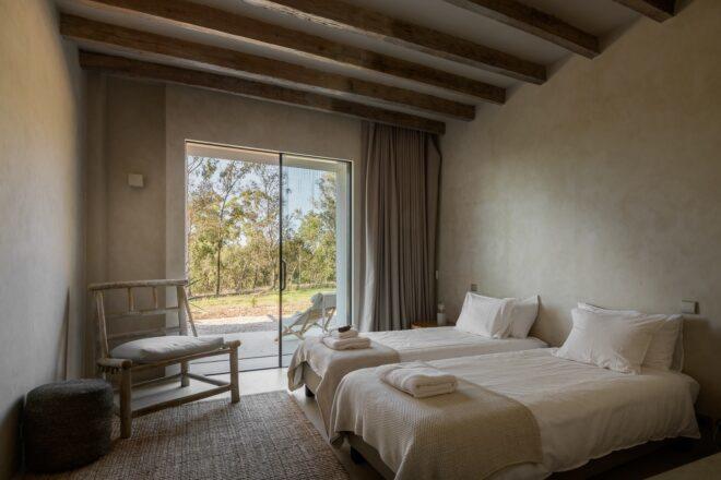 A bedroom with two beds, a chair, sliding glass doors and an exposed wood roof truss.