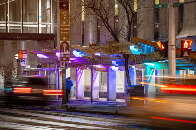 A cityscape with blurry cars passing in front of a wood canopy with blue and purple lights.