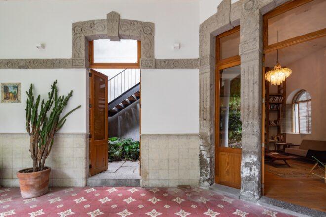 A hallway with vintage red tile flooring, a large potted plant and open doors leading to various parts of the building.