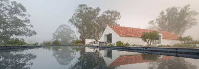 A foggy landscape with a large pool of water next to a simple A-line house with a red roof.