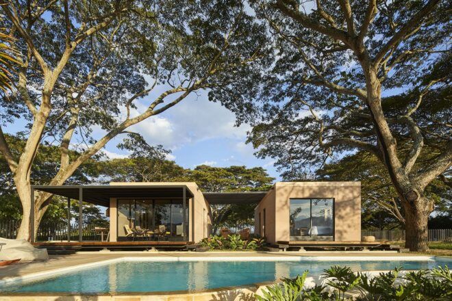 Two square volumes connected by a pergola. Large trees grow over the home and a pool sits in front of the home.
