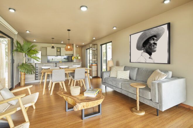 A living room with wood chairs and coffee table. A light blue/gray sofa sits on the right side of the room. Above the sofa is a black and white portrait photo.