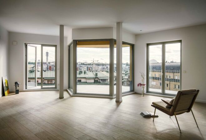 An open, mostly empty sitting room with white wall, light colored flooring and large windows.