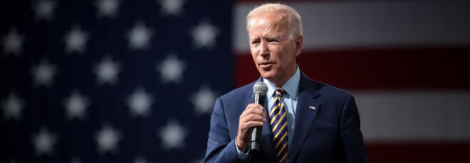 President Joe Biden holding a microphone while standing in front of an American flag background.