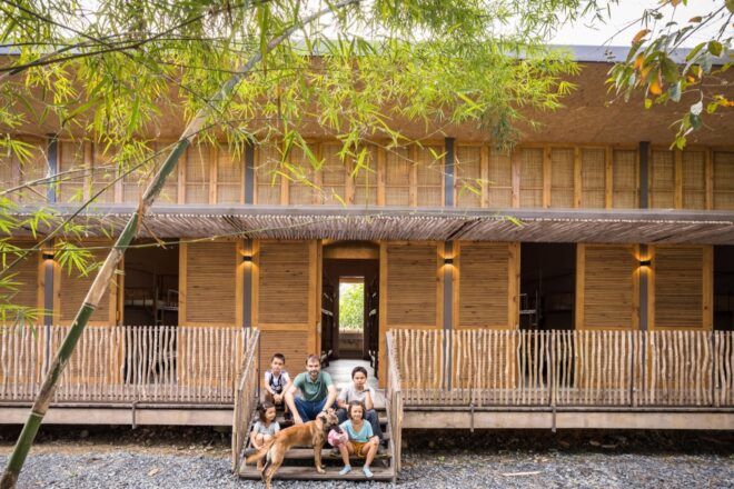 A two-story building with natural materials of bamboo and rice husk, with a family of five and a dog sitting on the front porch.
